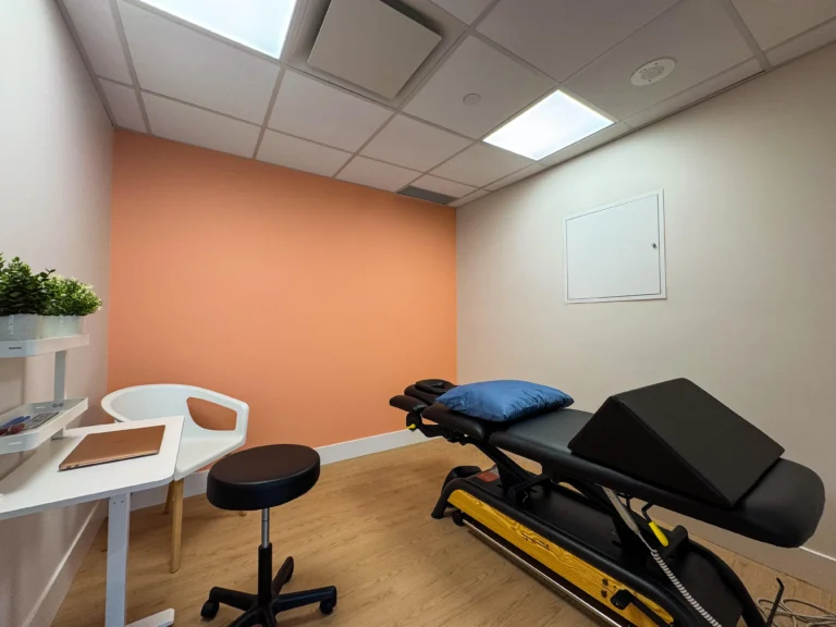 Private physio treatment and assessment room featuring a desk, a treatment table, a stool, and a chair, arranged for patient consultations and therapy sessions