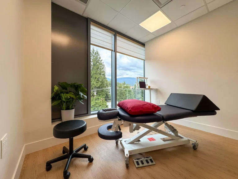 Multipurpose treatment room with a treatment table, work table, and stool, designed for various therapeutic and consultation uses, featuring a large window with a view of greenery.